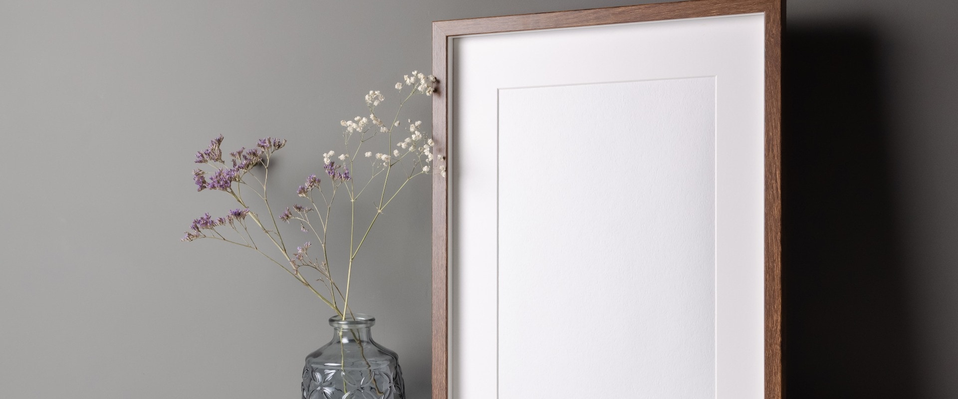 Vertical wooden frame mockup for artwork, photo and print presentation on white table over grey wall, dry flowers in vase.